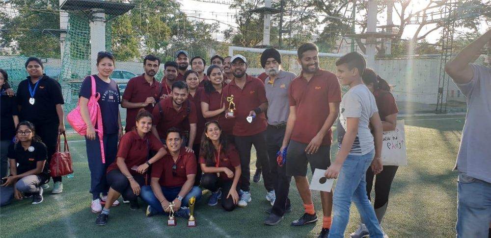 A smiling team in maroon shirts on a sports field, holding trophies